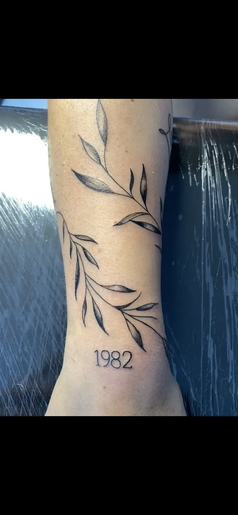 Fine line bladtak met het jaartal 1982 op de enkel, tattoo Gent