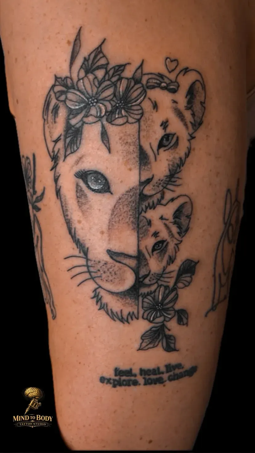Black and grey tattoo van een leeuwin met welp en bloemen op het bovenbeen, met lettering feel heal live explore love change, tattoo Gent